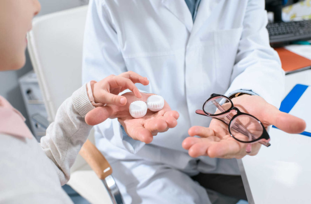 An optometrist holding a pair of glasses and a contact lens case in their hands while a young patient reaches to grab the contact lens case.