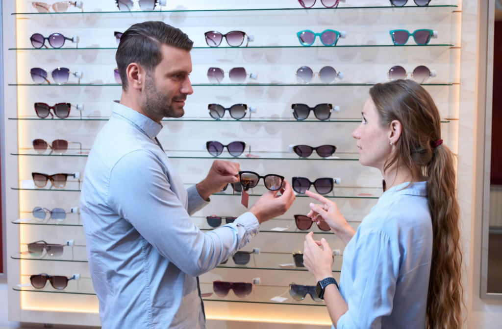 A young man holding up a pair of sunglasses and showing them to a young woman in front of a sunglasses display.