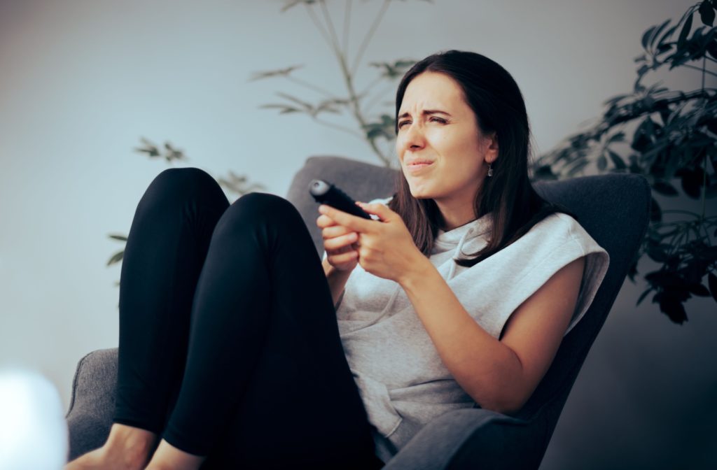 A woman sitting on a chair, squinting to see clearly on a TV while holding a remote.