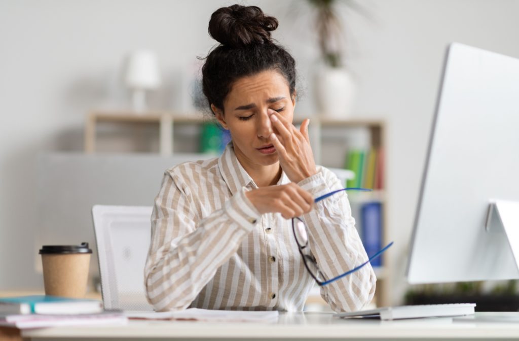 A persons suffering from stringy eye mucus while sitting at their desk rubbing their irritated eyes.