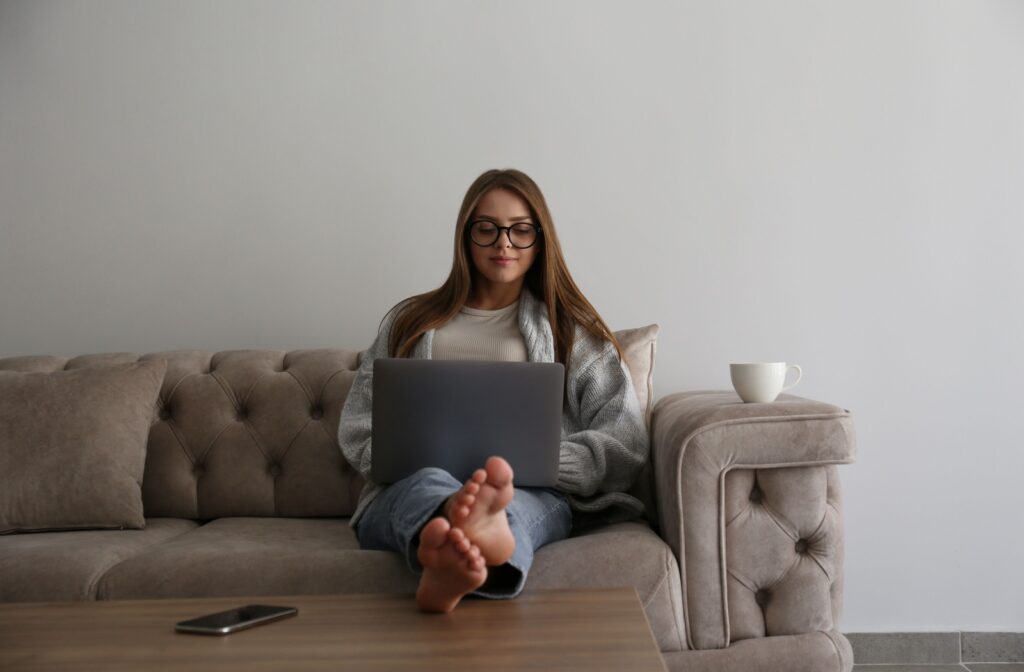 A person sitting on the coach, working on their laptop wearing blue light glasses.