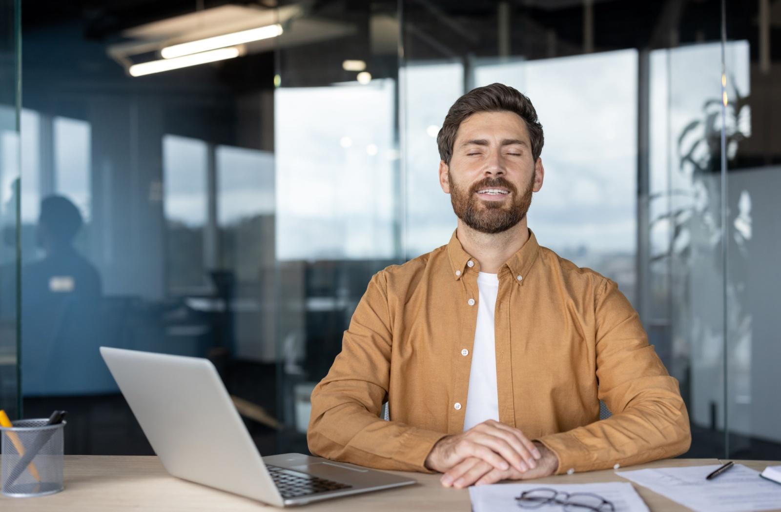 A person sitting in an office in front of a laptop, closing their eyes to give them a minute of rest from screen time.