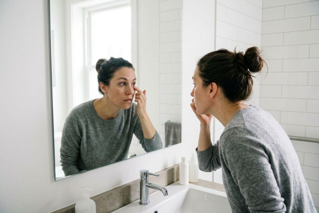 Person examining skin near their eye in a bathroom mirror, touching their face.